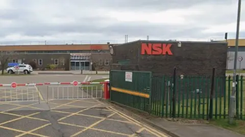 Google NSK Bearing's factory in Peterlee. A red-and-white raisable barrier blocks the road next to a brown-brick security hut. A sign shows the name of the company in red lettering. The factory stands behind it.