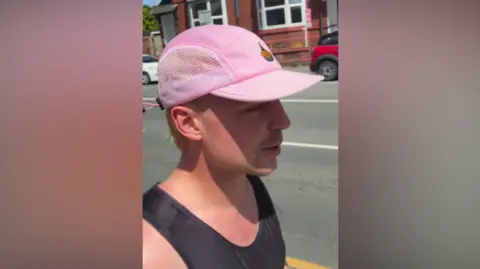 A man wearing black and a pink baseball cap is side on to the camera. A road is behind him, with parts of two cars and a building in the background.
