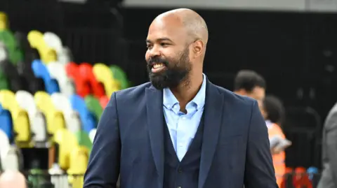 Gary Robinson A bald man with a beard wearing a three-piece navy suit. There are multi-coloured spectator seats behind him.