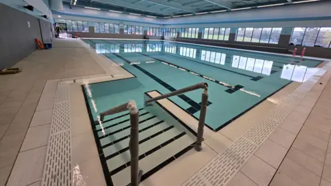 A large indoor swimming pool with turquoise water and dark lane markings on the tiled floor. Metal handrails lead down steps into the water in the foreground. The pool hall has floor-to-ceiling windows along two walls looking out onto trees and green space, with a worker in hi-vis visible at the far end.