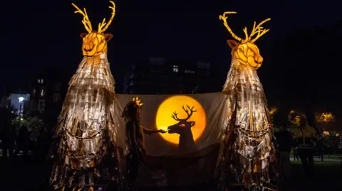 An installation featuring deer-like tall creatures which are glowing in an orange light. A person can be seen standing in front of a white sheet, feeding a lit up shadow of a deer.