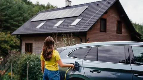 A woman in jeans and a yellow T-shirt charges her electric car, which is parked in front of a house with solar panels on the roof.