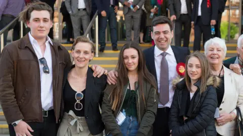 Getty Images The family look happy and are all dressed casually, standing in a row in front of the convention centre steps. Jimmy is very tall and wearing a brown leather jacket over a white shirt and black trousers, with a pair of sunglasses tucked into the neck of his shirt, and medium length, dark blonde hair held back by a hairband. Van Heel is wearing a tailored black jacket and black scoop-necked tshirt over beige paper-bag style trousers. Her dark blonde hair is worn medium length and slicked back in a quiff. Rosie wears a khaki green bomber jacket over a dark green t-shirt and blue jeans and has waist length, dark brown hair with bleached tips. Annie is wearing a black puffer jacket and a black top and has long, straight, dark blonde hair. Eileen is wearing a cream linen jacket over a black t-shirt and has ear length white hair worn in a pixie cut.
