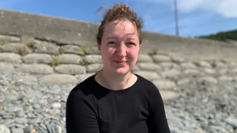 Poppy has curly dark hair tied back. She's wearing a plain black top and is sitting on a pebble beach with a stone wall behind her on a sunny day.