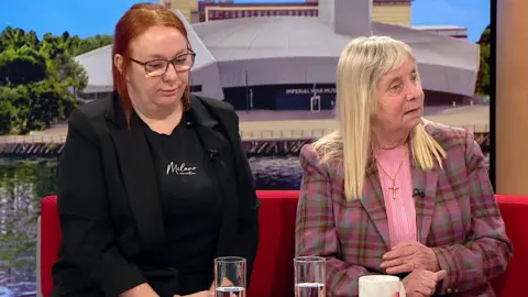 On the set of BBC Breakfast Charlotte Hennessy (L) has long red hair. dark glasses and is wearing a dark jacket and top while Margaret Aspinall (R) has long grey/blonde hair a tartan jacket with shades of brown and violet and a pink top. She wears a gold cross on a chain.