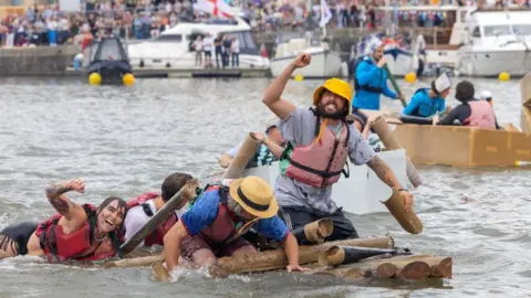 Bristol Harbour Festival An image of several people wearing hats, T-shirts and lifejackets clinging and sitting on three small boats made out of cardboard. A make-shift cardboard boat in the foreground appears to be sinking in the grey water. In the background can be seen a bridge lined with spectators.