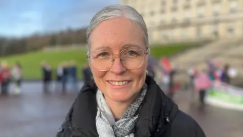 A woman with grey hair wearing large clear lensed glasses is pictured. She is wearing a black coat and a grey scarf and is smiling.