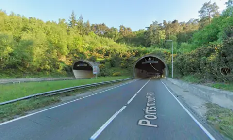 A stretch of road leading into a tunnel. It is a sunny day.