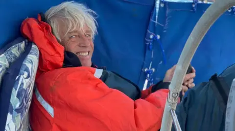 Facebook/Theros Sailing Adventure A smiling Brett Clibbery huddled on the deck of his yacht the Theros. He is wearing a red waterproof sailing jacket.