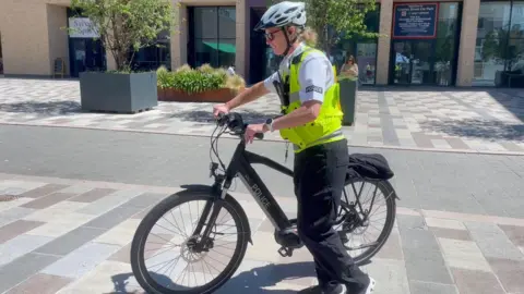 A police officer wearing a high=vis gilet steers a black police bicycle towards the left of the frame. She is wearing a white helmet, black trousers and a white shirt.