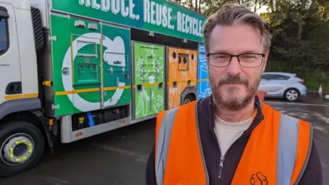 A councillor stands dressed in an orange high visibility jacket in front of one of Swindon Borough Council's waste disposal trucks