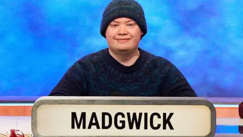 Kai Madgwick sits wearing a navy jumper and knitted navy hat looking at the camera with a smile 