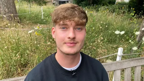 The image is a close-up outdoor portrait of a young man seated on a wooden bench. He has light brown wavy hair, blue eyes and light facial stubble, and is wearing a dark crew-neck sweatshirt over a white T-shirt. A small clip-on microphone is attached to the sweatshirt collar.