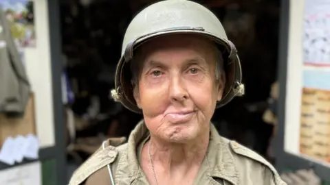 A close up of a man in khaki army fatigues and hard hat. His face is disfigured, with nose and mouth twisted to the right. The open doors of a shed can be seen behind him.