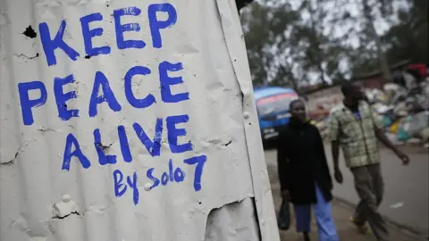 EPA Pedestrians walk in front of a wall with a message of peace painted recently by local street artist Solomon Muyundo, also known as Solo7, on its door in Kibera slum, one of the opposition leader Raila Odinga"s strongholds in the capital Nairobi, Kenya, 23 July 2017.