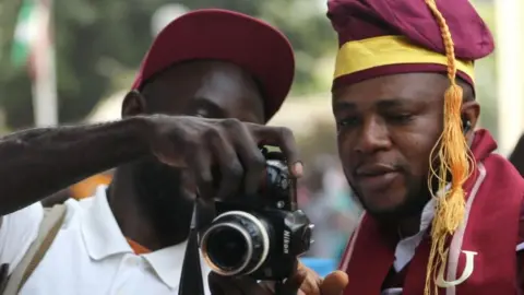 EPA A photographer shows photos on his camera for a graduating student outside the venue during 53rd convocation ceremony of the University of Lagos, Nigeria, 18 January 2023. The University of Lagos, one of the first generation universities in Nigeria, celebrates its 60th anniversary with 53rd convocation ceremony graduating two octogenarian students for graduate studies.