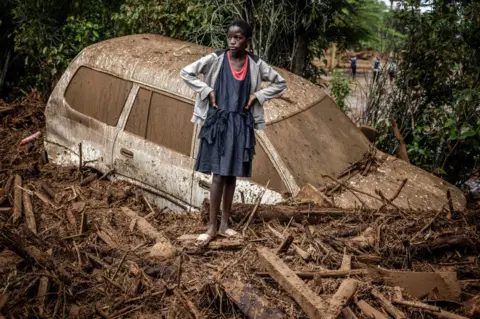 LUIS TATO/AFP A girl looks on next to a damaged car buried in mud in an area heavily affected by torrential rains and flash floods in the village of Kamuchiri, near Mai Mahiu, on April 29, 2024.