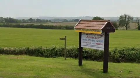 The image shows a wooden sign for Muston Grange Caravan Park, with a wooden roof over the top. There is a small wooden direction sign next to it and rolling fields behind it, with trees and hedges in the background and a hedge in the foreground. 