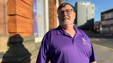 Rob Constantin/BBC Stan Robinson, standing outside a building, wearing a purple top, with his chin up, looking at the camera. He has on black glasses, has short hair and a grey beard.