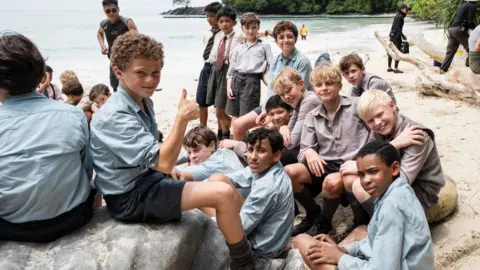 A group of young boys are sat down on a beach on a remote island. They are posing at the camera. One boy is sitting on a rock, putting his thumb up at the camera.