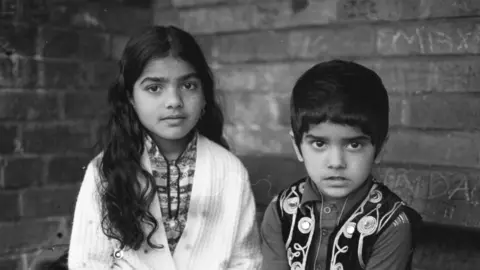 Sharon Hepburn Two children sit side by side against a brick wall, looking toward the camera.
