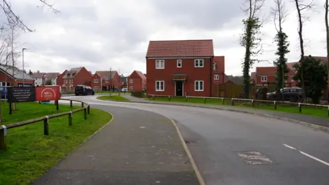 A new build red brick two-storey detached house, standing on the bend of a road, with low level fencing in front of grass verges. Other houses are in the background.