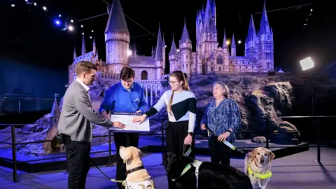 Warner Bros Studio/Guide Dogs A man and woman are feeling a raised map of the Hogwarts miniature model. The actual model towers behind them. They are holding leads attached to guide dogs. Another woman is stood to the side also with a guide dog. A member of staff wearing a blue uniform is holding the raised map.