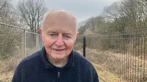 Peter Twentyman smiles stood next to fencing to protect the Manchester, Bolton and Bury Canal. A channel can be seen over a bank with trees on the other side.