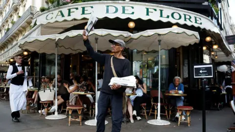 Reuters Ali Akbar, wearing a gray flat cap and a black dress, with a paper high in his right hand in front of the Cafe de Flore in Paris