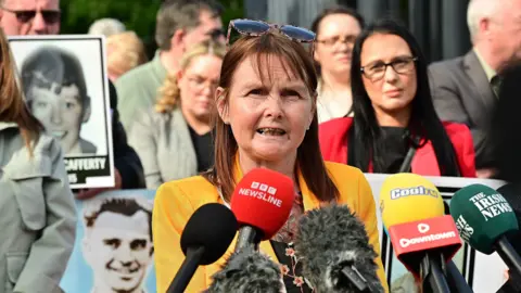 Pacemaker Jacqueline Butler speaking in front of microphones. She has brown hair, wearing a yellow jacket.