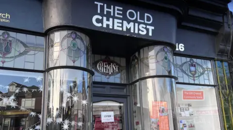 The front of an old building with stained glass and rounded windows. 
It is labelled The Old Chemist and has a sign in the window saying Community space