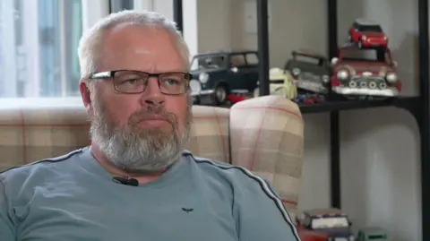 A man, Steve Lewsey, sat in an armchair in a blue t-shirt. He has a beard and glasses.