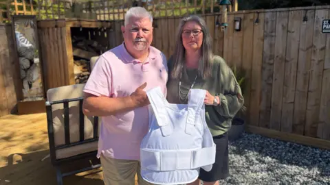 Claire Lewis is standing next to her partner Ged Mulheran holding a stab vest in front of them. They are in a garden with a fence behind them and both are wearing shorts and are in their 60s. She has ash blonde long hair, he is wearing a pink shirt and has a goaty beard and silver hair.