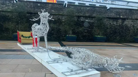 a christmas reindeer decoration lies on its side in Derry's Guildhall Square. It is broken off its plinth. A identical reindeer remains standing. Both are displayed to pull a sleigh that can be seen in the background in front of the city's walls decoration