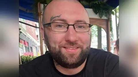 Head and shoulders photo of Jonathan Bruce, a man with a brown beard, black t-shirt and glasses, sitting outdoors at a restaurant