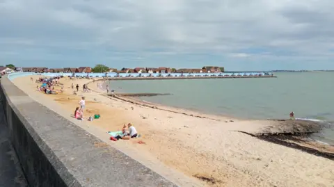 Canvey Island's beach, with the photo taken from behind the concrete sea wall. Adults and children are on the beach. The blue painted sea wall, facing out to sea, can be seen curving round the bay. It is slightly overcast above.