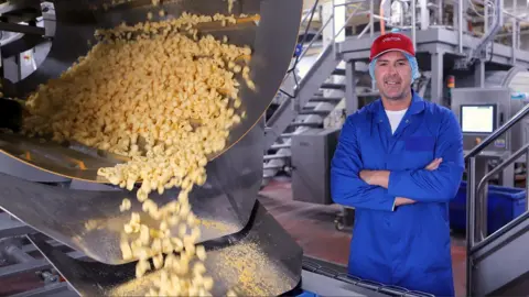 Paddy McGuinness standing on the production line wearing blue overall and a red cap. The curls can be seen cascading from a drum.