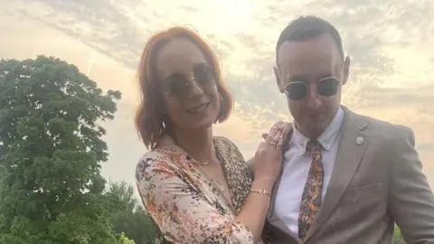 Family photograh Liv (left) and Sam Shaw standing on a golf club house balcony overlooking the golf course. Liv has chestnut coloured hair and is wearing a brown beige and orange patterned dress and has her arms resting on Sam's shoulder. He has short brown hair and is wearing a beige suit with paisley patterned tie. They are both wearing sunglasses. It is a sunny day. 