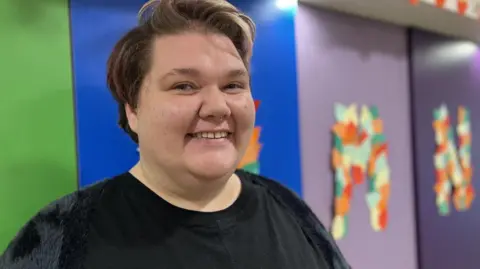 A person with short hair - coloured light brown with flecks of pink - and a small silver nose ring smiles at the camera. They are wearing a black top. The background is colourful with purple, blue and green blocks and large hand-made letter murals in autumnal colours.