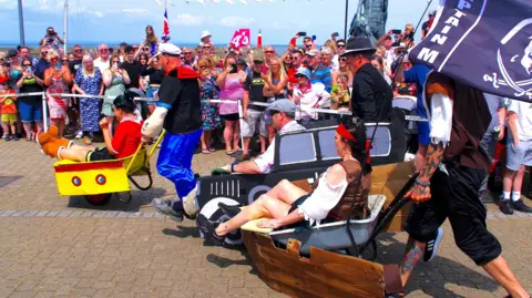 Watchet Wheelbarrow Race Three different sets of competitors take part in the Watchet wheelbarrow races, with the people in the wheelbarrows wearing different types of fancy dress. It is a sunny day, and dozens of people are watching from the roadside