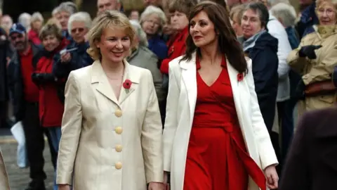 PA Media/John Giles Kathryn Apanowicz smiles while walking past crowds of people. She is wearing a cream-coloured coat with a poppy pinned to it. She is walking alongside Carol Vorderman, who is wearing a red dress and cream-coloured coat as well as a poppy.