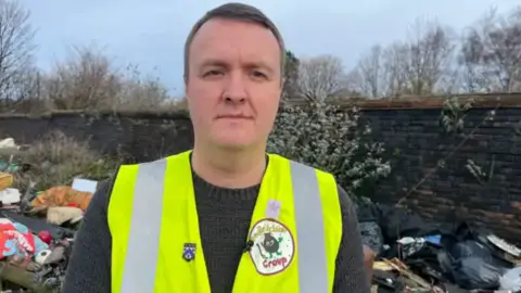 Brennan stands for the photo in front of a pile of fly-tipped rubbish behind. He's wearing a hi-vis jacket and brown jumper.