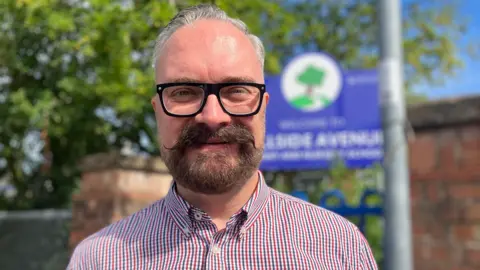 A man with glasses and a handlebar moustache and beard smiles towards the camera outside a school on a sunny day