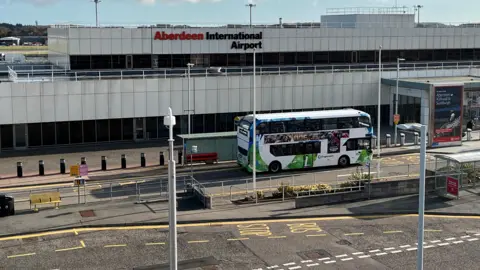 A wide shot of the front of Aberdeen International Airport's main terminal building, with a double decker bus parked outside