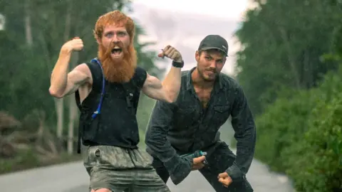 Two men pose, showing off their biceps, in the middle of a road in the jungle.