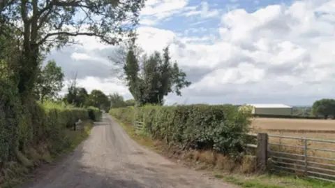 A country lane, with a metal gate into a field on the right, and a large barn can be seen in the distance