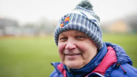 Darrell Williams smiling at the camera with the blurred rugby pitch in the background. He is wearing a blue and white bobble hat and a navy and red coat. 