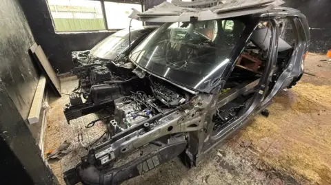 A grey car shell with no doors, wheels or side windows. The car looks like it is ready to be scrapped.