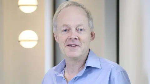 Professor Hugh Markus, a man who is standing in front of a white wall. He is looking directly at the camera and smiling and is wearing a pale blue shirt.