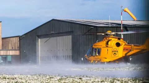 The helicopter is either landing or taking off outside a hanger next to a frosty grass. A wind sock can be seen in the background.